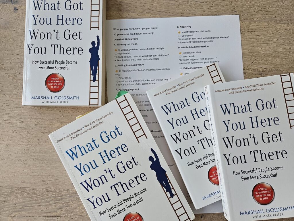 Multiple copies of the book 'What Got You Here Won’t Get You There' by Marshall Goldsmith spread on a wooden table, featuring a ladder motif along the spine.