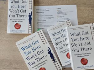 Multiple copies of the book 'What Got You Here Won’t Get You There' by Marshall Goldsmith spread on a wooden table, featuring a ladder motif along the spine.