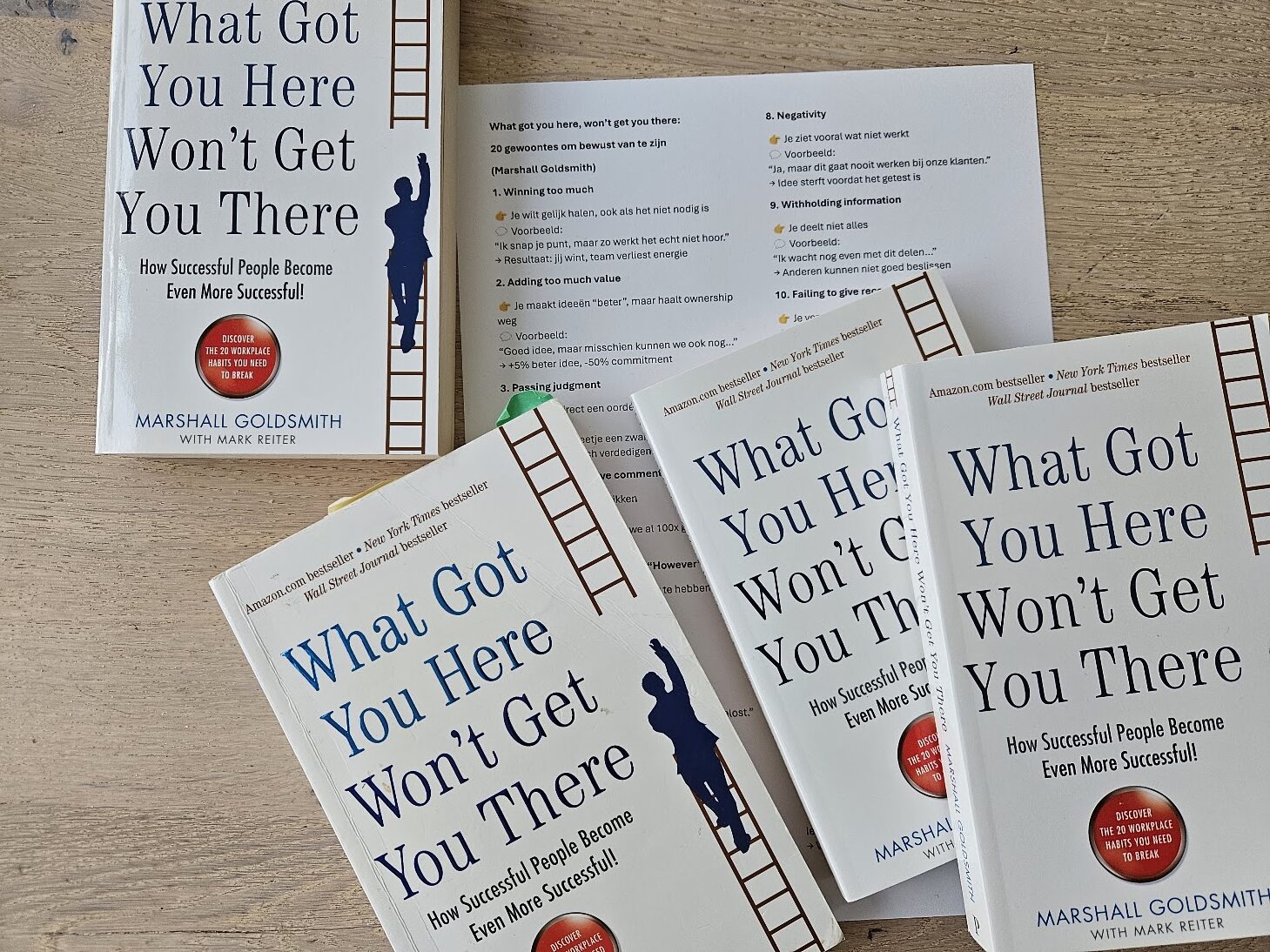 Multiple copies of the book 'What Got You Here Won’t Get You There' by Marshall Goldsmith spread on a wooden table, featuring a ladder motif along the spine.