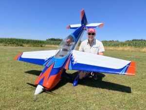 Man kneeling beside a large blue, orange, and white remote-controlled airplane on a grassy airfield, smiling at the camera.