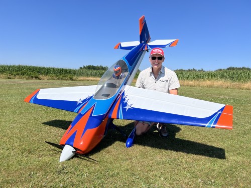 Jeroen met Extra 104inch 500x375 - VOLK.NL - VOLK.NL Man kneeling beside a large blue, orange, and white remote-controlled airplane on a grassy airfield, smiling at the camera.